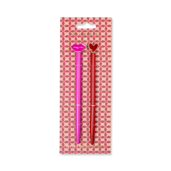 Two pens, one pink with lips at the top and one red with a heart at the top, on a pink checked cardboard backing.
