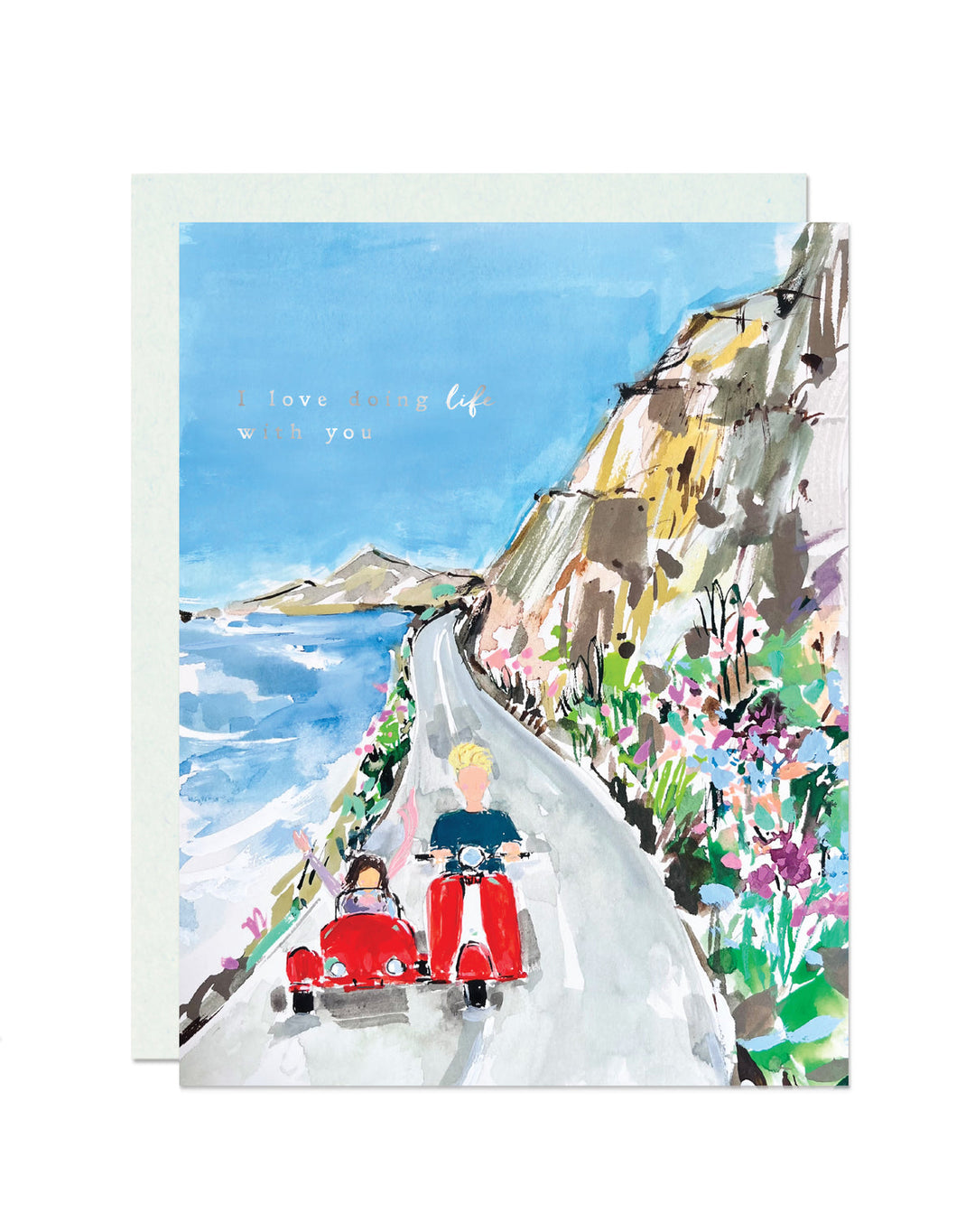 A watercolor-style greeting card with a light blue envelope behind it. The card shows a coastal road scene with a person riding a red scooter and another person in a red sidecar, with the text 'I love living life with you' printed near the top.