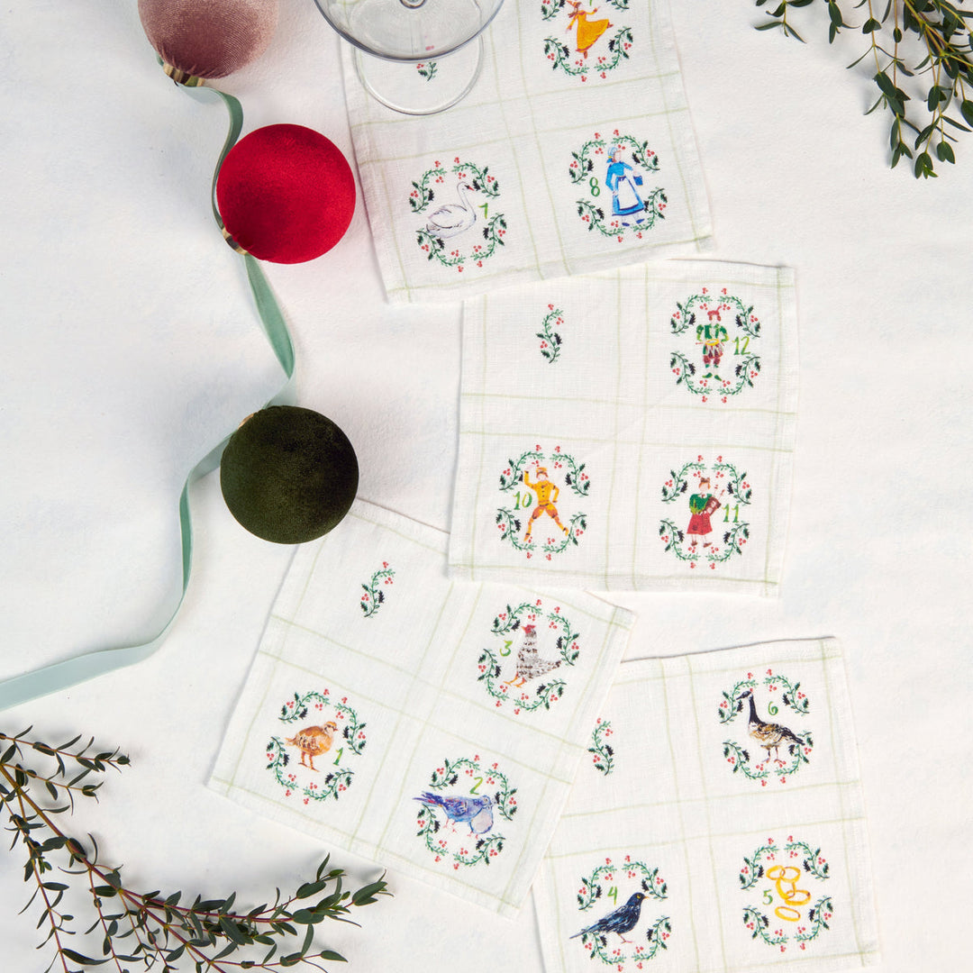 A styled flat-lay of multiple white square napkins with embroidered multicolor  motifs from the 12 days of Christams, arranged on a white surface with holiday ornaments, ribbon, and greenery.
