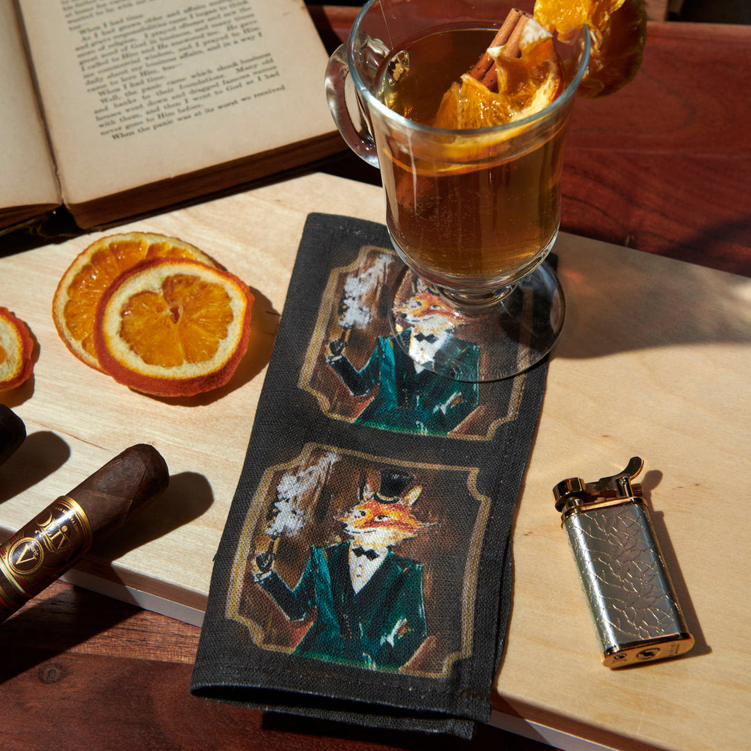 Dark cocktail napkin with drawing of a fox in a tuxedo with a cocktail and a lighter and a cigar and some orange slices