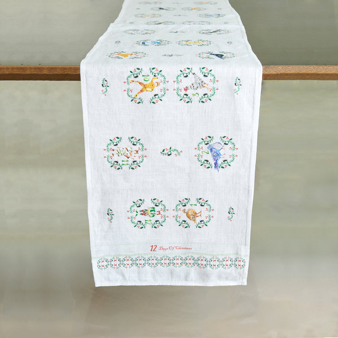 A white linen table runner with colorful embroidered motifs of the gifts for the 12 days of Christmas draped over a table edge.
