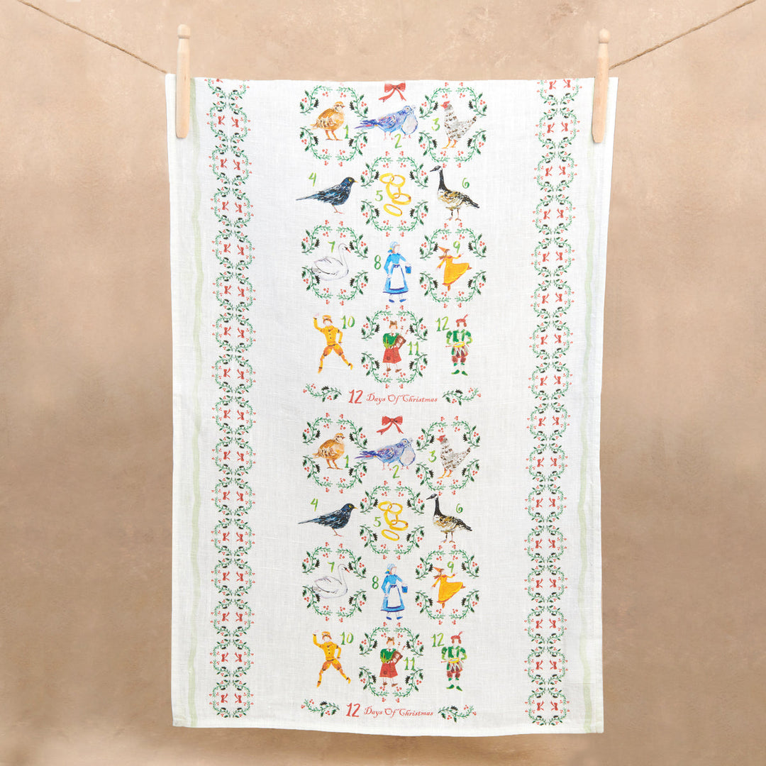 A white linen tea towel with figures of the 12 gifts for the 12 days of Christmas pinned to a clothesline.