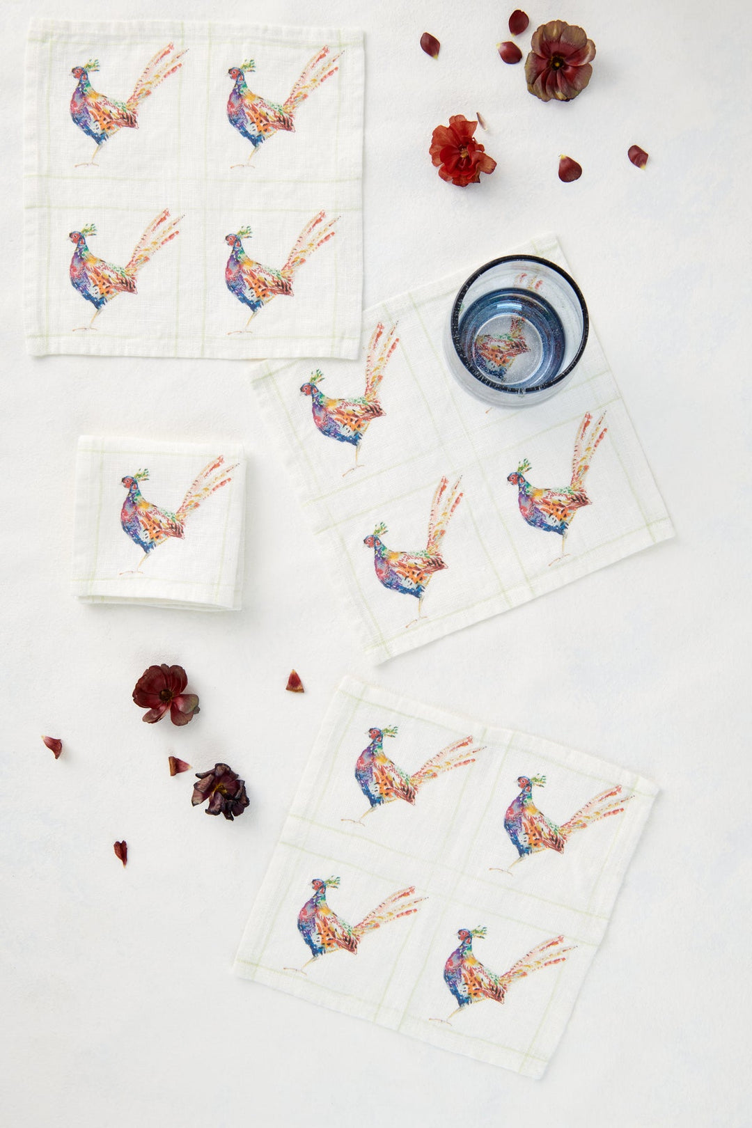 Several white linen napkins featuring embroidered colorful pheasant birds, arranged on a white background with scattered red flower petals and a small blue-rimmed glass.
