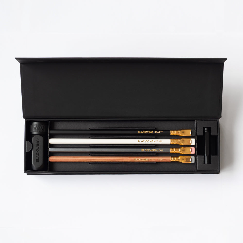 The hinged lid of the black box is open to show a black pencil sharpener, four different pencils and a black point guard.