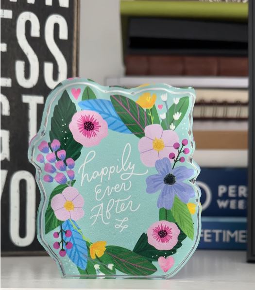 Decorative irregularly shaped plaque with floral design and 'happily ever after' text in the center, in front of a stack of books.