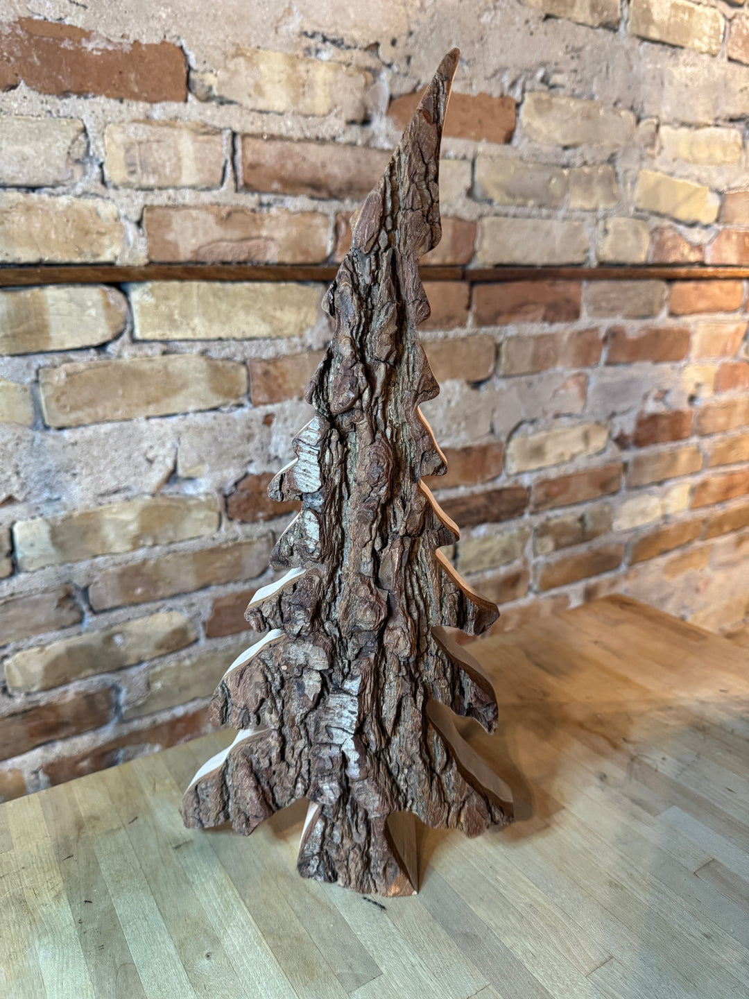 Carved wooden Christmas tree with a combination of bark and smooth wood, with a conical shape and detailed trunk, in front of a brick wall