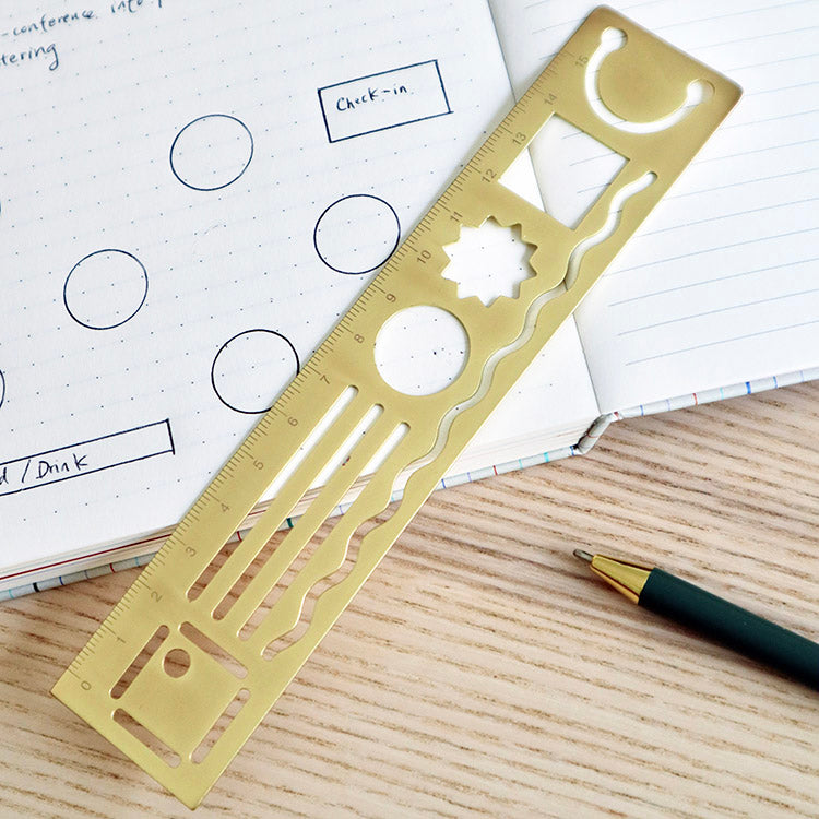 Brass 15" ruler with template of geometric shapes resting on a notebook showing shapes drawn with the template.