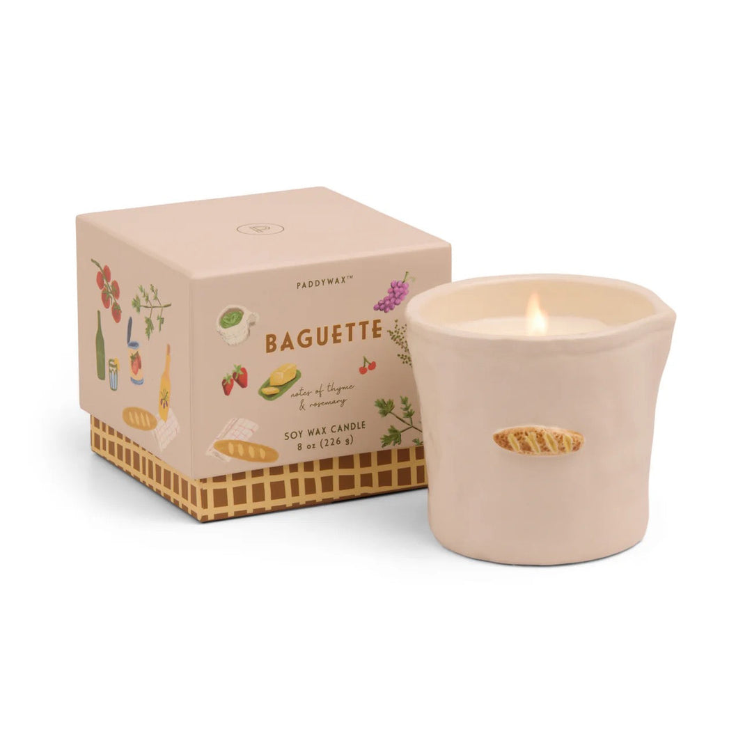 Burning candle in a beige ceramic vessel decorated with a single baguette, next to a gift box with images of bread, cheese, grapes, strawberries and cherries.