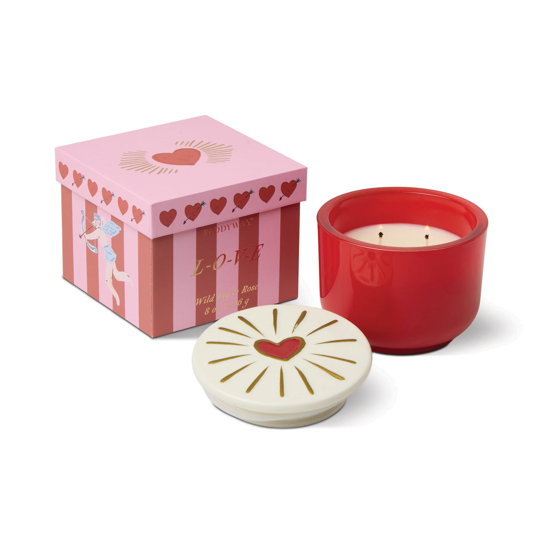 Scented candle in red container with a white lid featuring a heart design, next to a pink gift box with heart patterns and cupid.