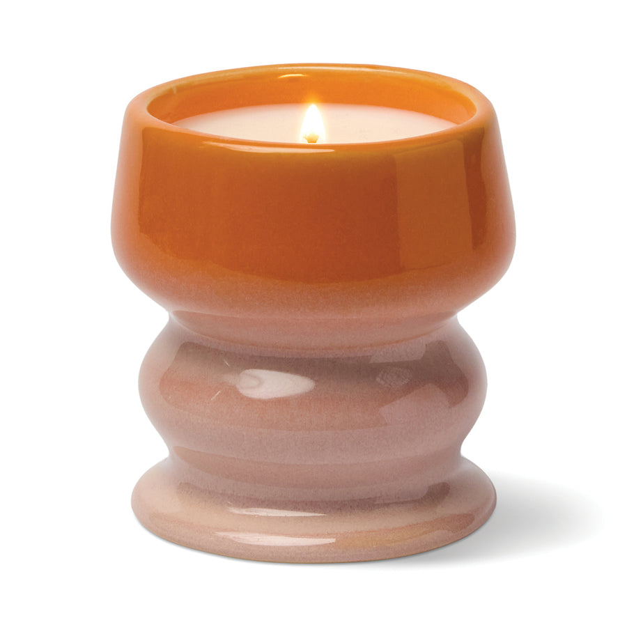 Lit candle in a curvaceous ceramic vessel in colors fading from off orange to beige.