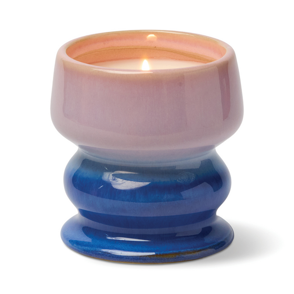 Lit candle in a curvaceous ceramic vessel in colors fading from pink to blue 