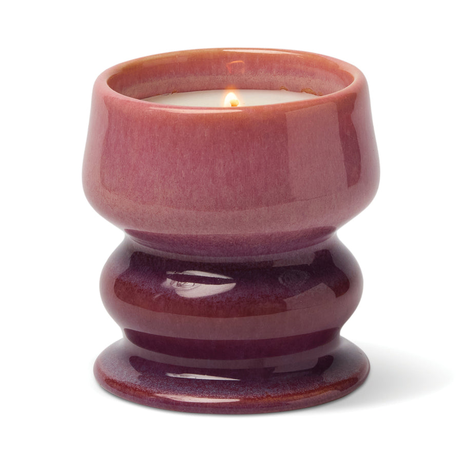 Lit candle in a curvaceous ceramic vessel in colors fading from off light burgundy to dark burgundy.