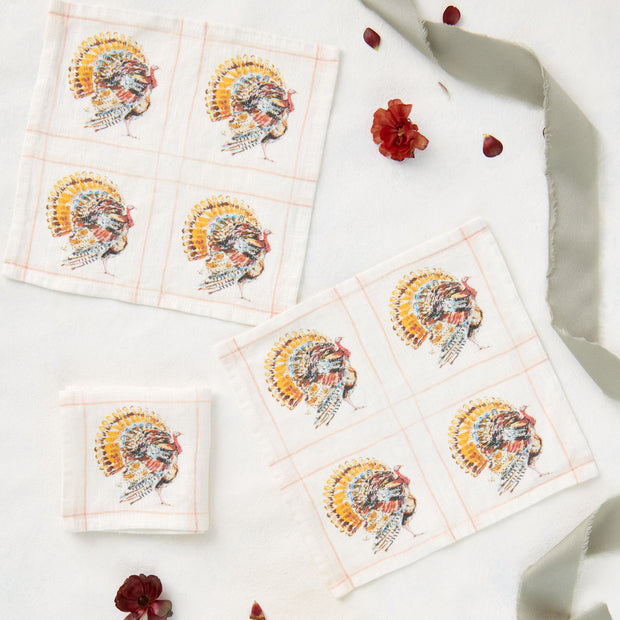 Three square cotton napkins with a Thanksgiving turkey print, arranged with decorative red flowers and a gray ribbon.