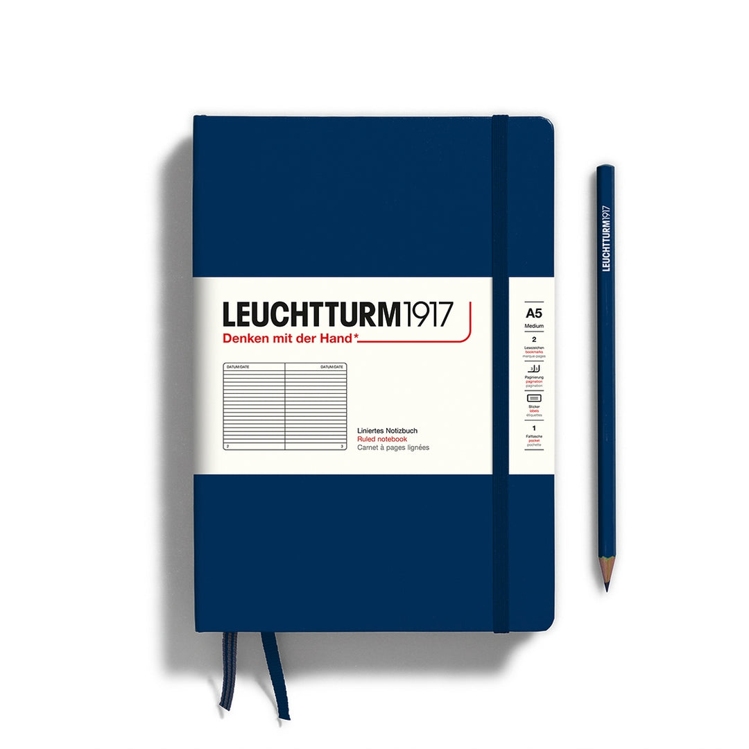A navy Leuchtturm1917 A5 ruled page pocket notebook with a white front label and navy elastic closure,  next to a navy Leuchtturm1917 pencil.