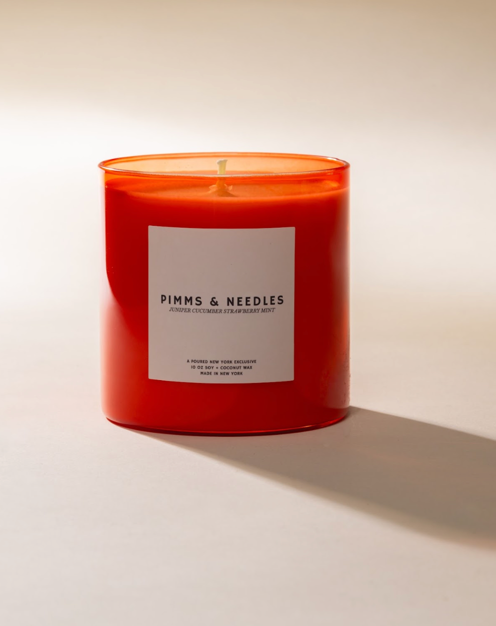 Candle in orange glass jar with white label having the text 'PIMMS & NEEDLES'.