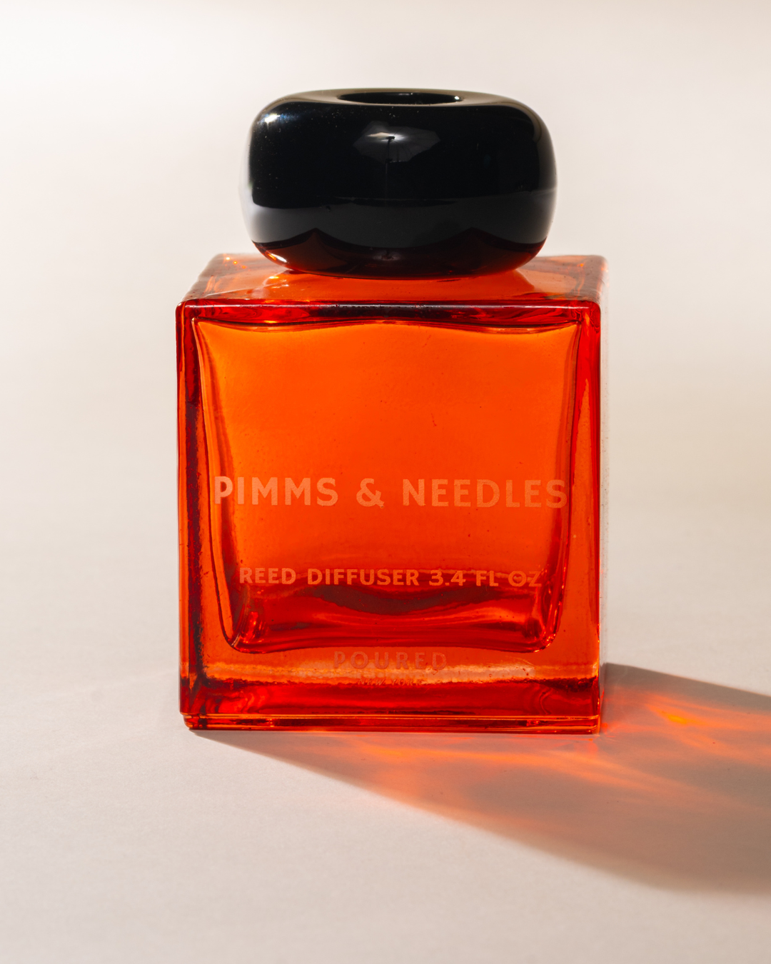 A square glass reed diffuser bottle with vibrant orange liquid and a glossy black cap, featuring the brand name 'PIMMS & NEEDLES' printed on the front.
