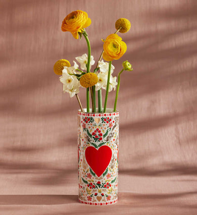 Decorative vase with a red heart and floral patterns holding yellow and white flowers.