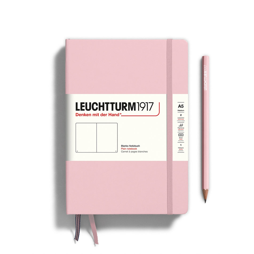 A Leuchtturm1917 A5 hardcover plain page notebook in powder pink with a white label on the front, shown closed with an elastic closure and ribbon bookmark. A matching powder pink pencil is placed vertically to the right.