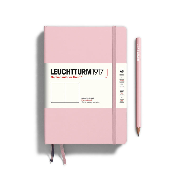 A Leuchtturm1917 A5 hardcover plain page notebook in powder pink with a white label on the front, shown closed with an elastic closure and ribbon bookmark. A matching powder pink pencil is placed vertically to the right.