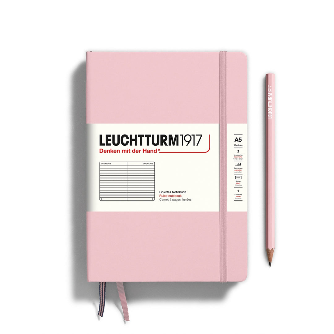 A Leuchtturm1917 A5 hardcover ruled page notebook in powder pink with a white label on the front, shown closed with an elastic closure and ribbon bookmark. A matching powder pink pencil is placed vertically to the right.