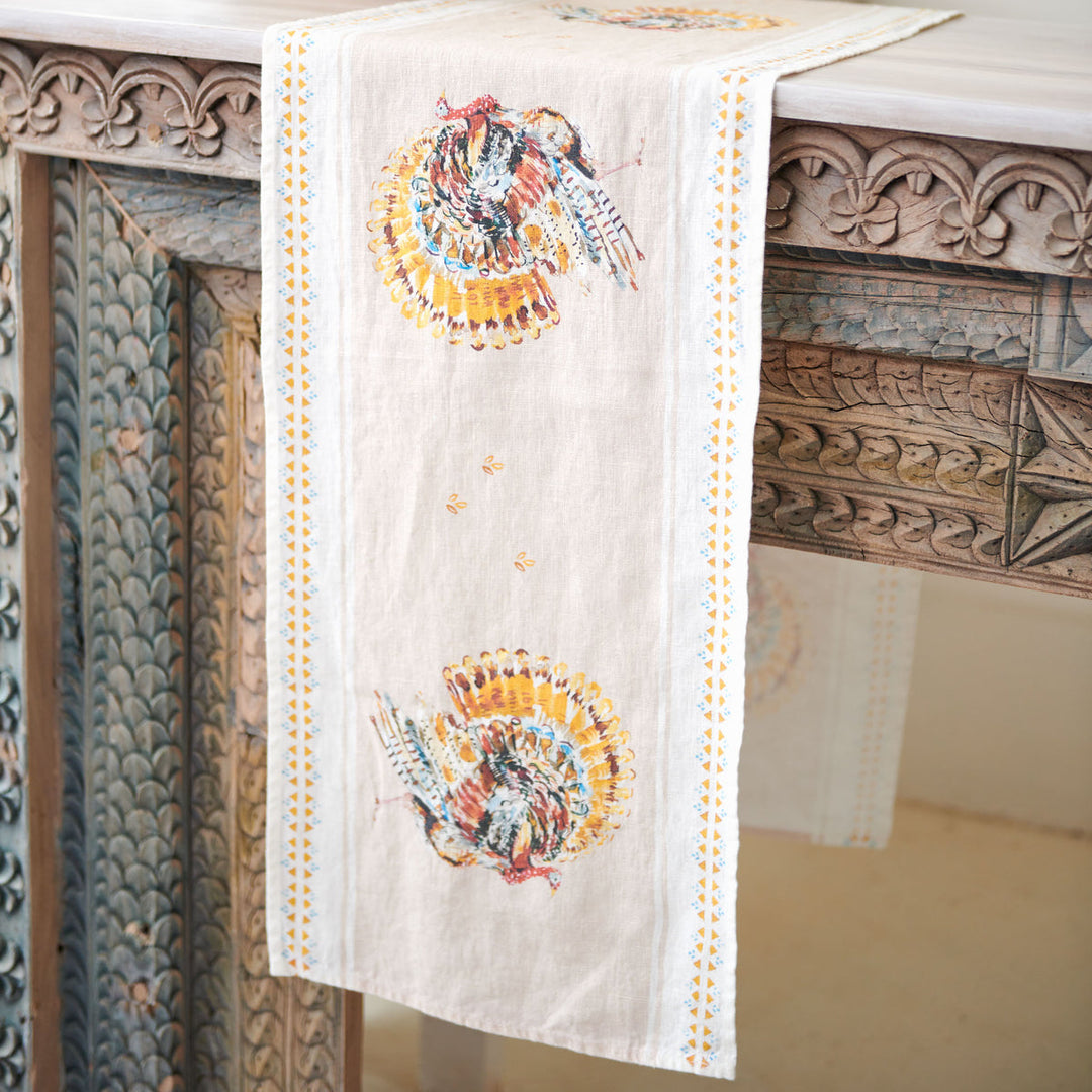 An off-white table runner with turkeys and a dotted border, draped over an ornate carved wooden table.