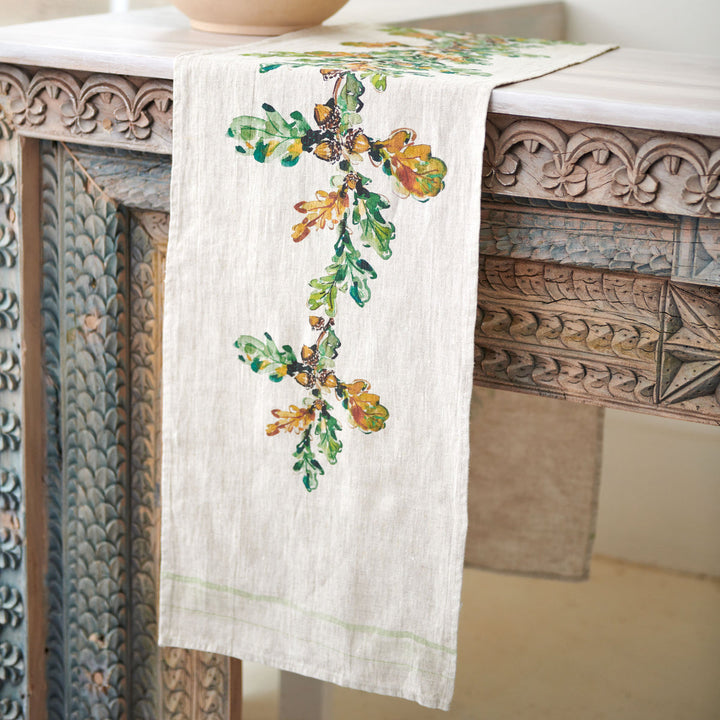 One end of an off-white linen table runner draped over an ornate carved wooden table, with a design of oak leaves with acorns running down the center.
