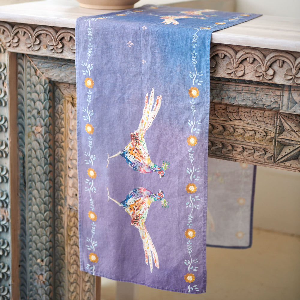 One end of a blue table runner draped over an ornate carved wooden table showing a pair of pheasants facing each other, with a border of flower heads.