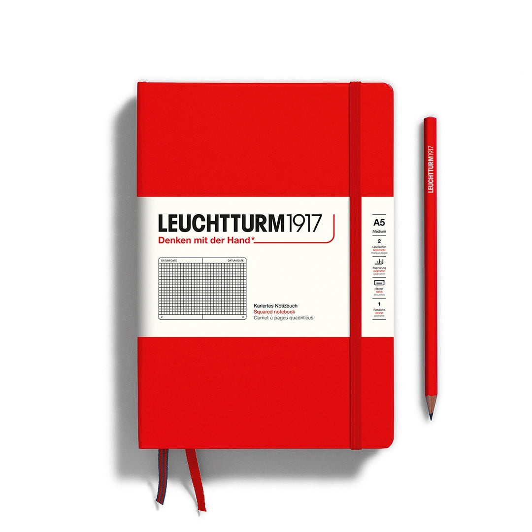 A Leuchtturm1917 A5 hardcover squared page notebook in red with a white label on the front, shown closed with an elastic closure and ribbon bookmark. A matching red pencil is placed vertically to the right.