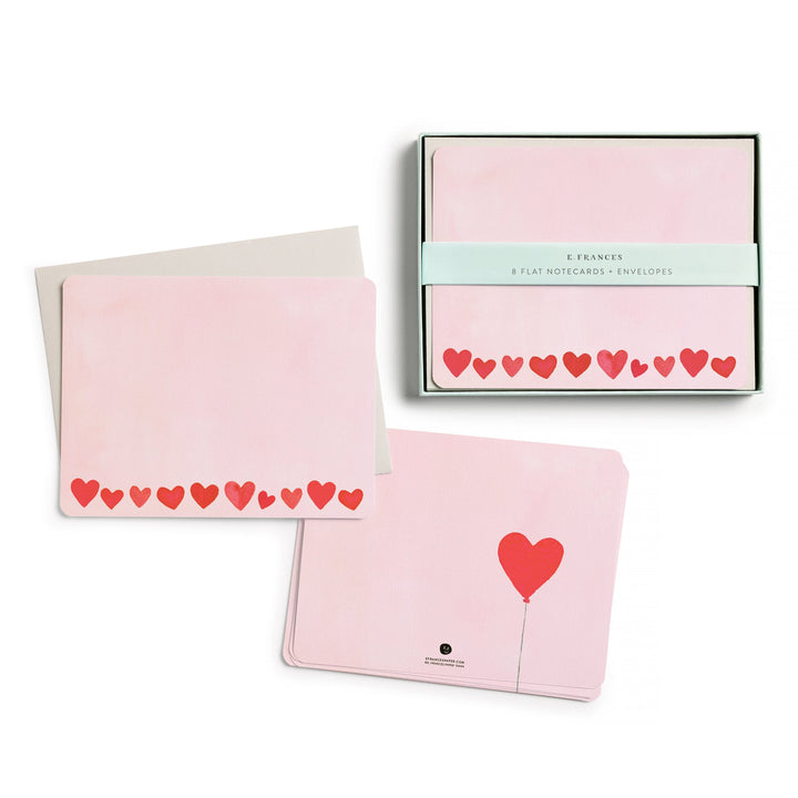 The box of cards, with belly band, next to two stacks of cards, one showing the front, with red hearts across the bottom, and the other the back, with a single red heart on a string.