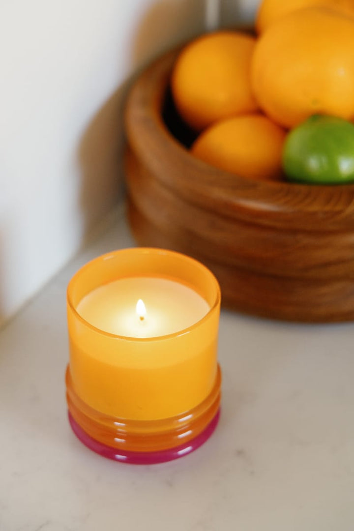 Lit candle in orange glass container with red lid used as a base underneath, net to a wooden bowl of fruit.