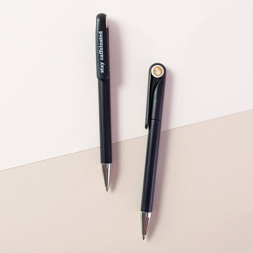 Two sides of a black retractable ballpoint pen with silver tips. One side shows the word 'stay caffeinated' iin white lowercase letters, and the other side has a circular cap insert with an image of a coffee  plant.