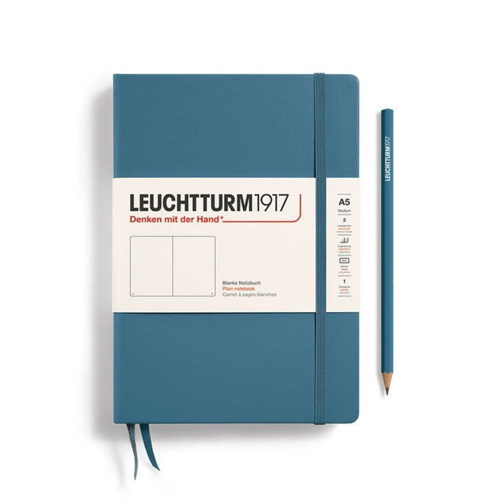 A Leuchtturm1917 A5 hardcover plain page notebook in stone blue with a white label on the front, shown closed with an elastic closure and ribbon bookmark. A matching stone blue pencil is placed vertically to the right.