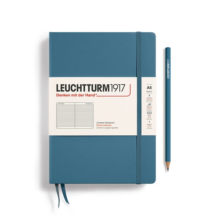 A Leuchtturm1917 A5 hardcover ruled page notebook in stone blue with a white label on the front, shown closed with an elastic closure and ribbon bookmark. A matching stone blue pencil is placed vertically to the right.