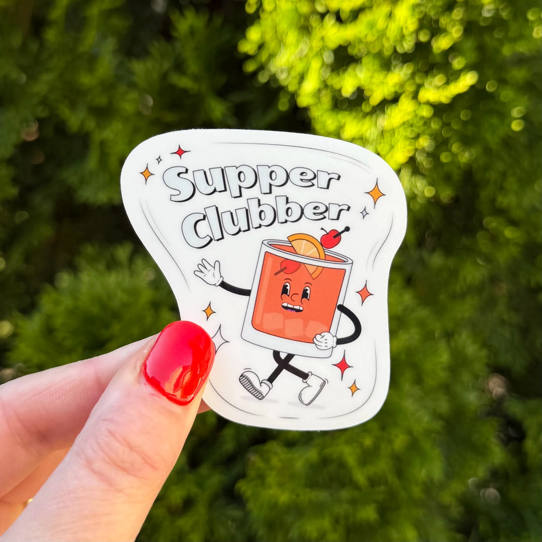 A hand with bright red nail polish holds a die-cut sticker featuring a brandy old fashioned as a cartoon character with the text 'Supper Clubber'.