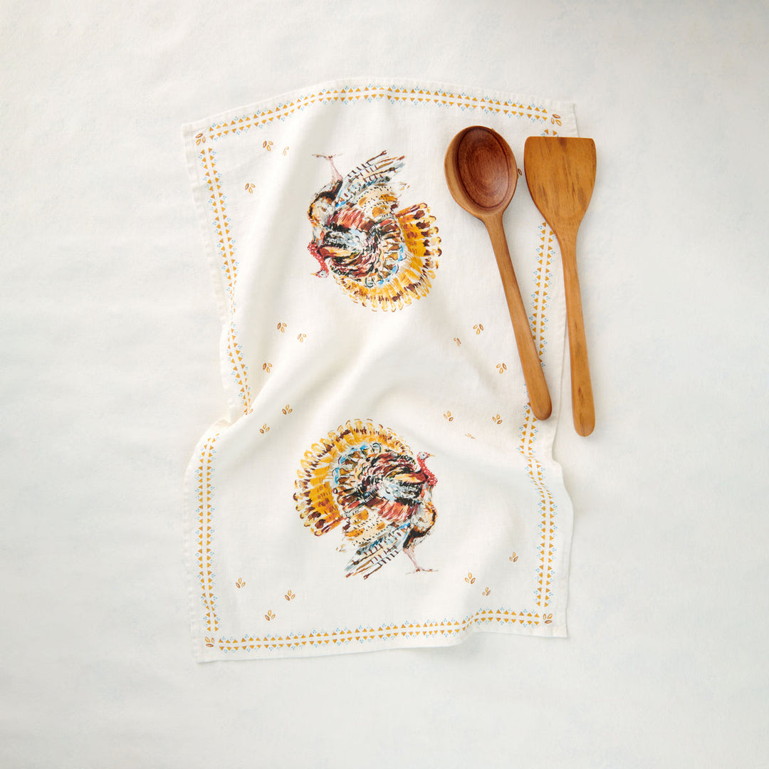 An white linen kitchen towel with a painted turkeys and a border of a double row of golden specks. Wooden serving spoon and spatula lie on the towel.