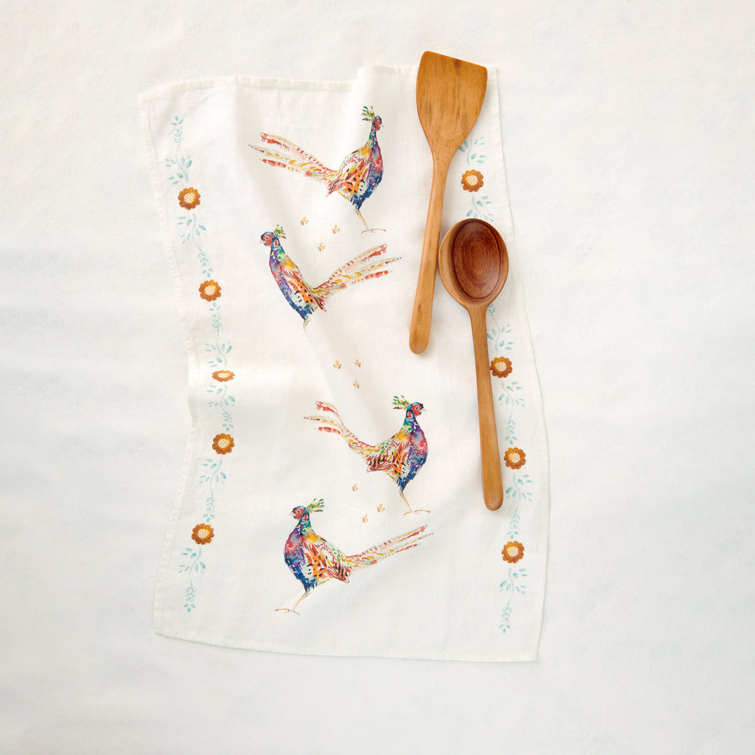 An white linen kitchen towel with a painted pheasants and a border of fall flower buds. Wooden serving spoon and spatula lie on the towel.