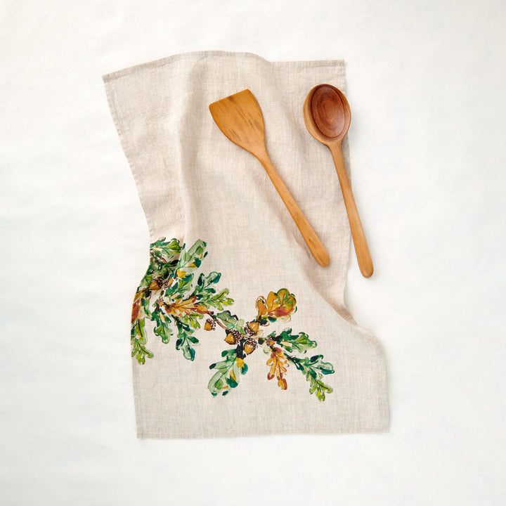 An off-white linen kitchen towel with a painted green leafy branch with acorns. Wooden serving spoon and spatula lie on the towel.