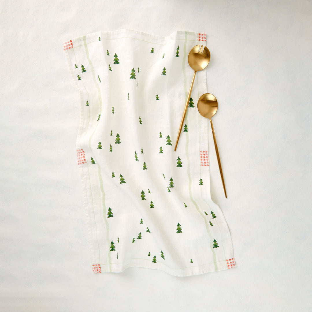 A white cotton tea towel with a green pine tree pattern and small red checkered corner accents. Two gold spoons rest on the towel.