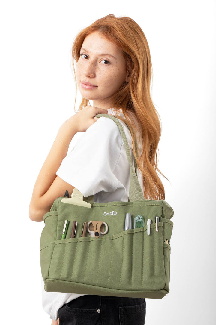 Woman with long red hair has the green studio bag over her shoulder. The narrow pockets contains scissors, pens and other narrow art supplies.
