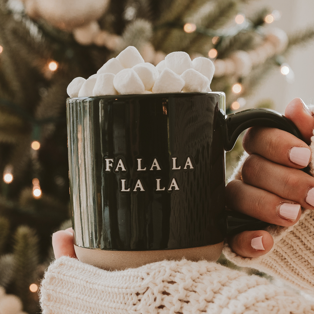 Black mug with 'FA LA LA LA LA' text, filled with hot chocolate and marshmallows, against a festive background.