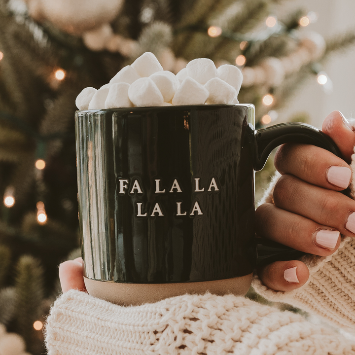 Black mug with 'FA LA LA LA LA' text, filled with hot chocolate and marshmallows, against a festive background.