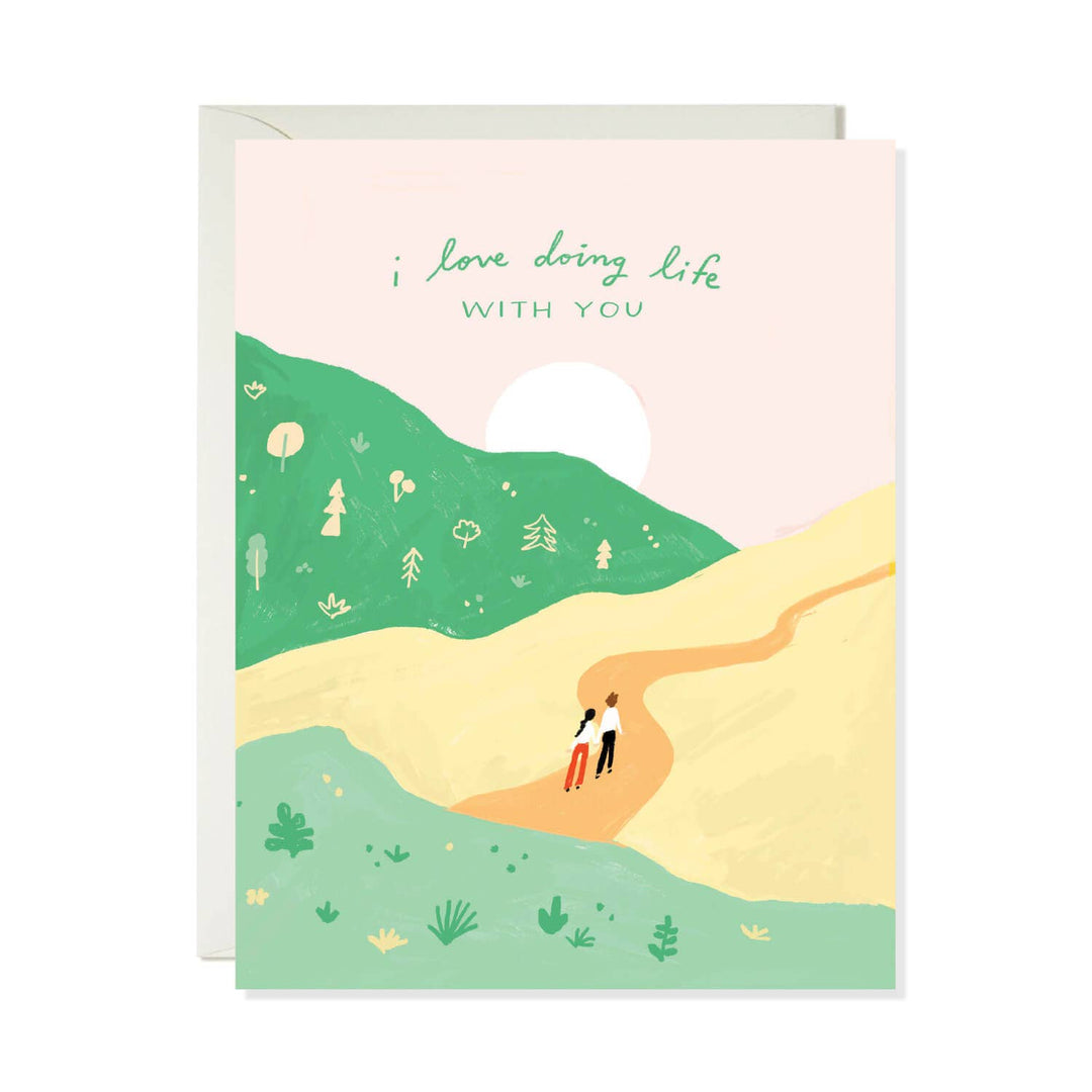 A greeting card with a light pink envelope behind it. The card features an illustration of a person walking along a path through green hills and a pale yellow field under a soft pink sky, with the text 'i love doing life WITH YOU' printed at the top.