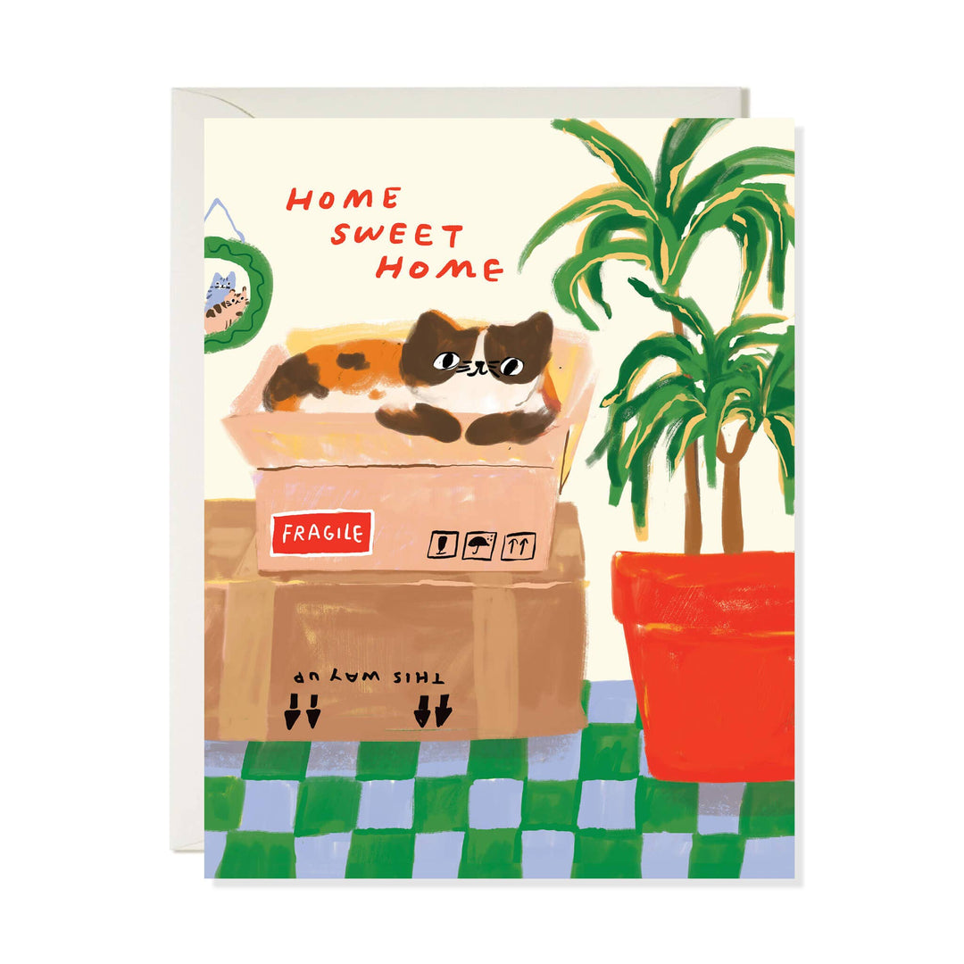 A greeting card with a whimsical illustration of a calico cat lounging in a cardboard box next to a potted palm, with the text 'HOME SWEET HOME'.