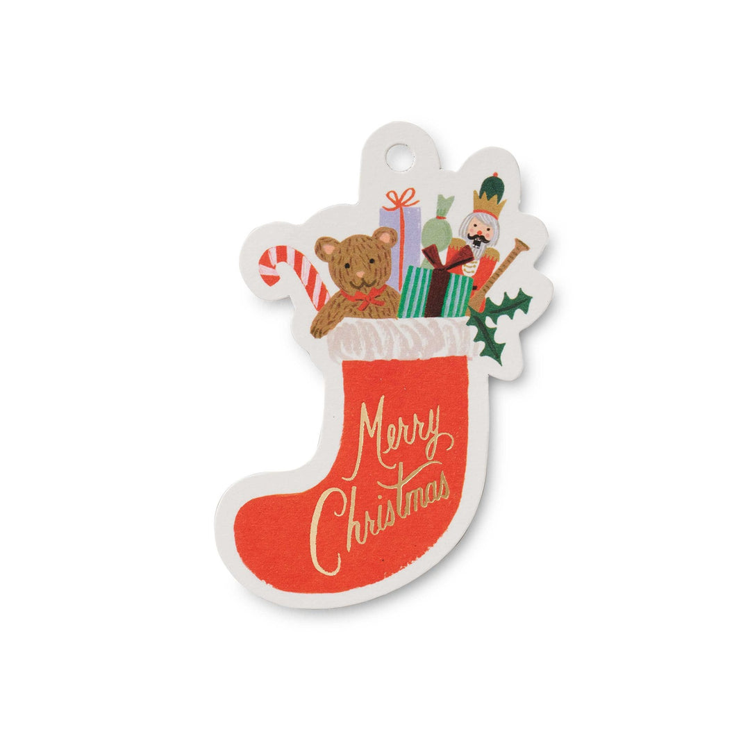 A Christmas stocking-shaped tag with a white border, featuring a red stocking with gold 'Merry Christmas' script, a teddy bear, candy cane, gifts, nutcracker, and holly on a white background.