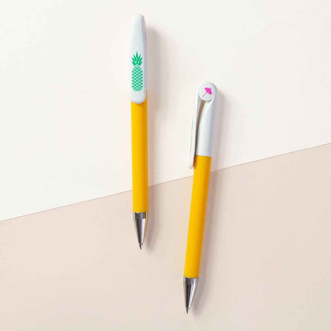 Two sides of a retractable ballpoint pen with a yellow barrel and white top and silver tips. One side has a green pineapple graphic, and the other side has a circular cap insert with an image of a drink umbrella.
