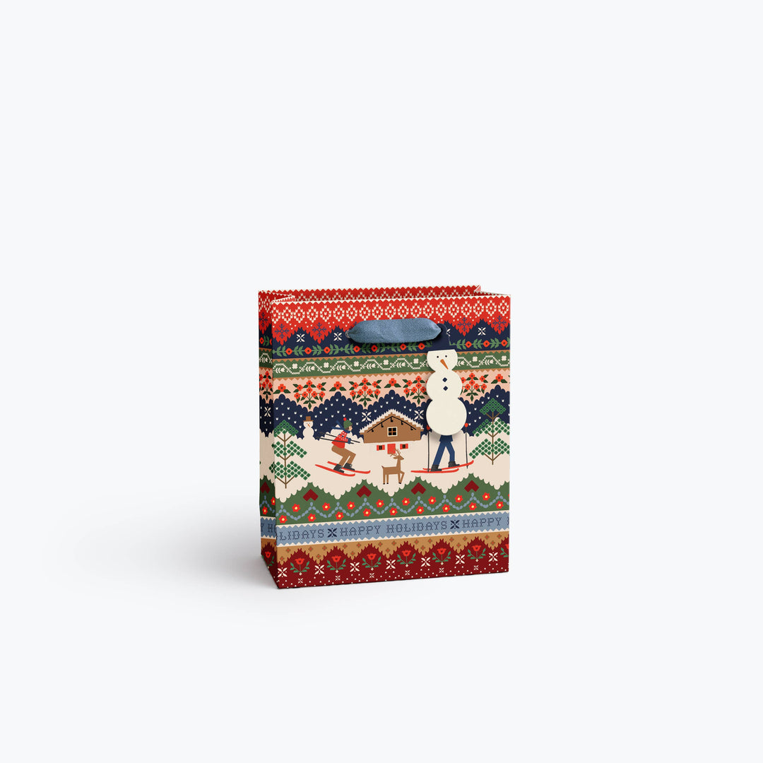 A medium gift bag with a colorful winter folk pattern featuring skiers, a cabin, a reindeer, with geometric borders in red, green, blue, beige and black, a gray fabric handle and snowman gift tag.