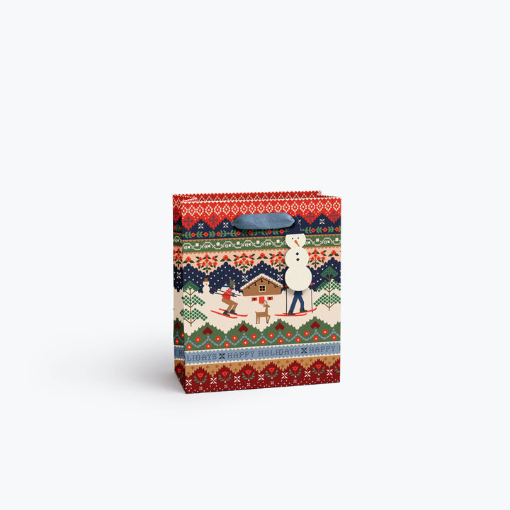A medium gift bag with a colorful winter folk pattern featuring skiers, a cabin, a reindeer, with geometric borders in red, green, blue, beige and black, a gray fabric handle and snowman gift tag.