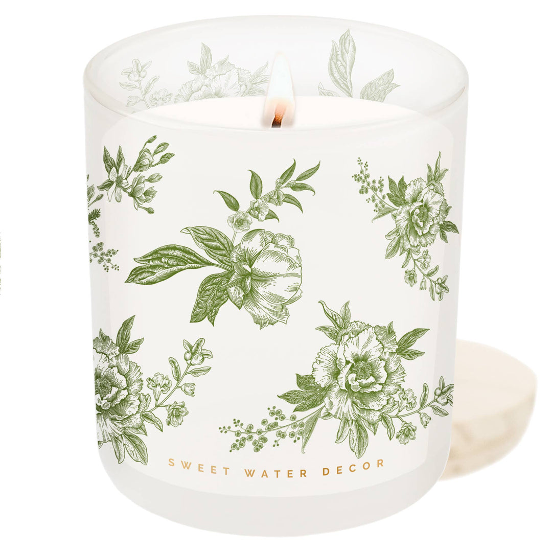 Lit candle in white opaque container with green delft pattern next to its wooden lid.