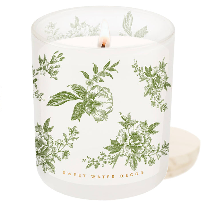 Lit candle in white opaque container with green delft pattern next to its wooden lid.
