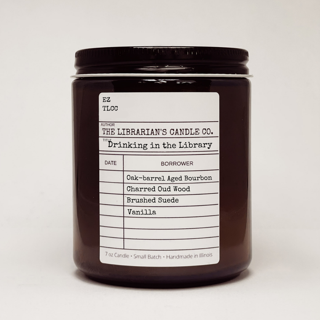 Candle jar with a label in the design of a library due date slip for the book "Drinking in the Library" detailing the candles scent notes: Oak-barrel Aged Bourbon, Charred Oud Wood, Brushed Suede, Vanilla.
